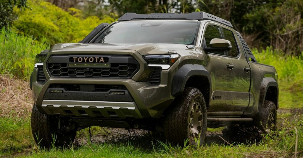 A green 2024 Toyota Tacoma pickup truck is driving on a dirt road surrounded by trees. The truck has a roof rack and a bed cover. The Toyota logo is visible on the front of the truck.