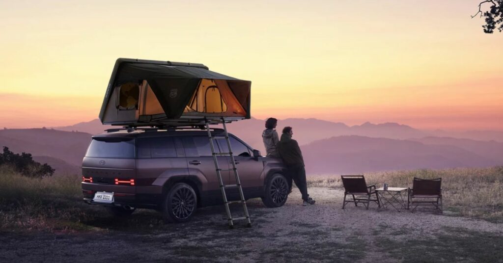 A brown SUV with a rooftop tent open, two people standing beside it at sunset.
