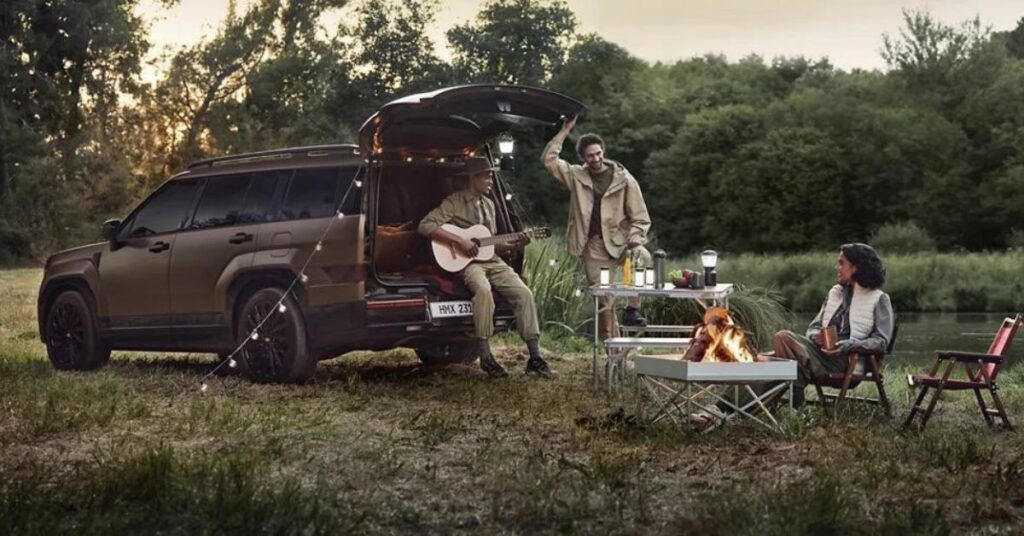 Three people camping with a brown SUV, man playing guitar, fire pit in the foreground.